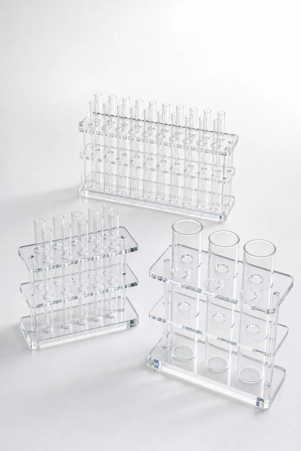 Clear acrylic (PMMA) laboratory tube racks with two drilled plates holding rows of empty test tubes in three hole sizes on a white bench