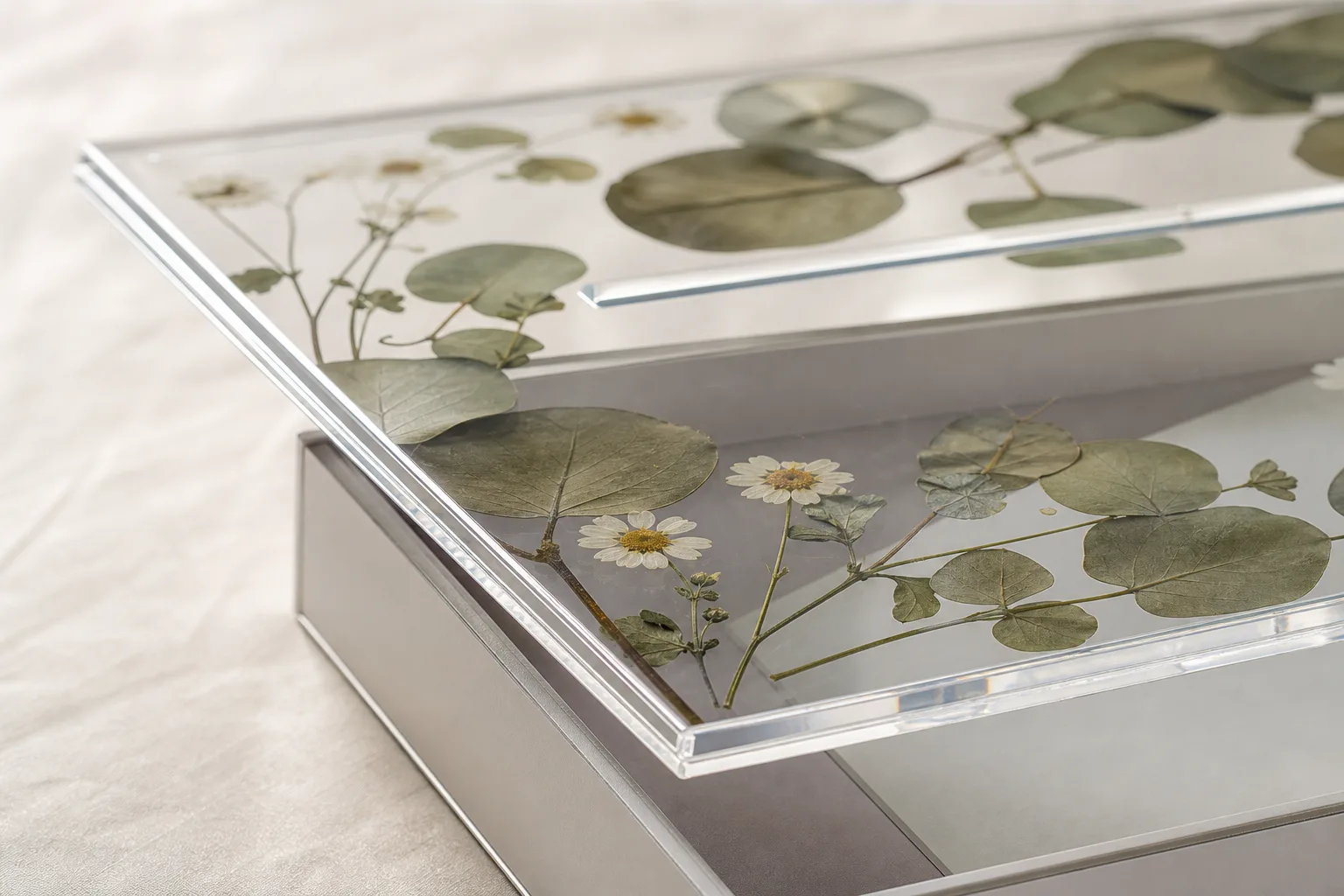 Clear acrylic wedding card box with pressed flowers laminated flat inside the lid panel, engraved names on the front face, and an envelope slot on top, styled on a linen-covered reception table
