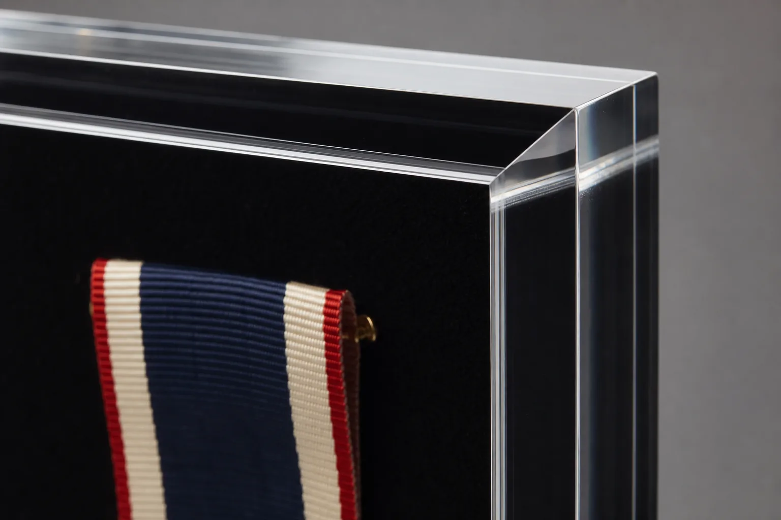 Close-up of a clear acrylic shadow box frame corner showing the polished front panel edge, deep recess, and black velvet backing with a pinned medal ribbon