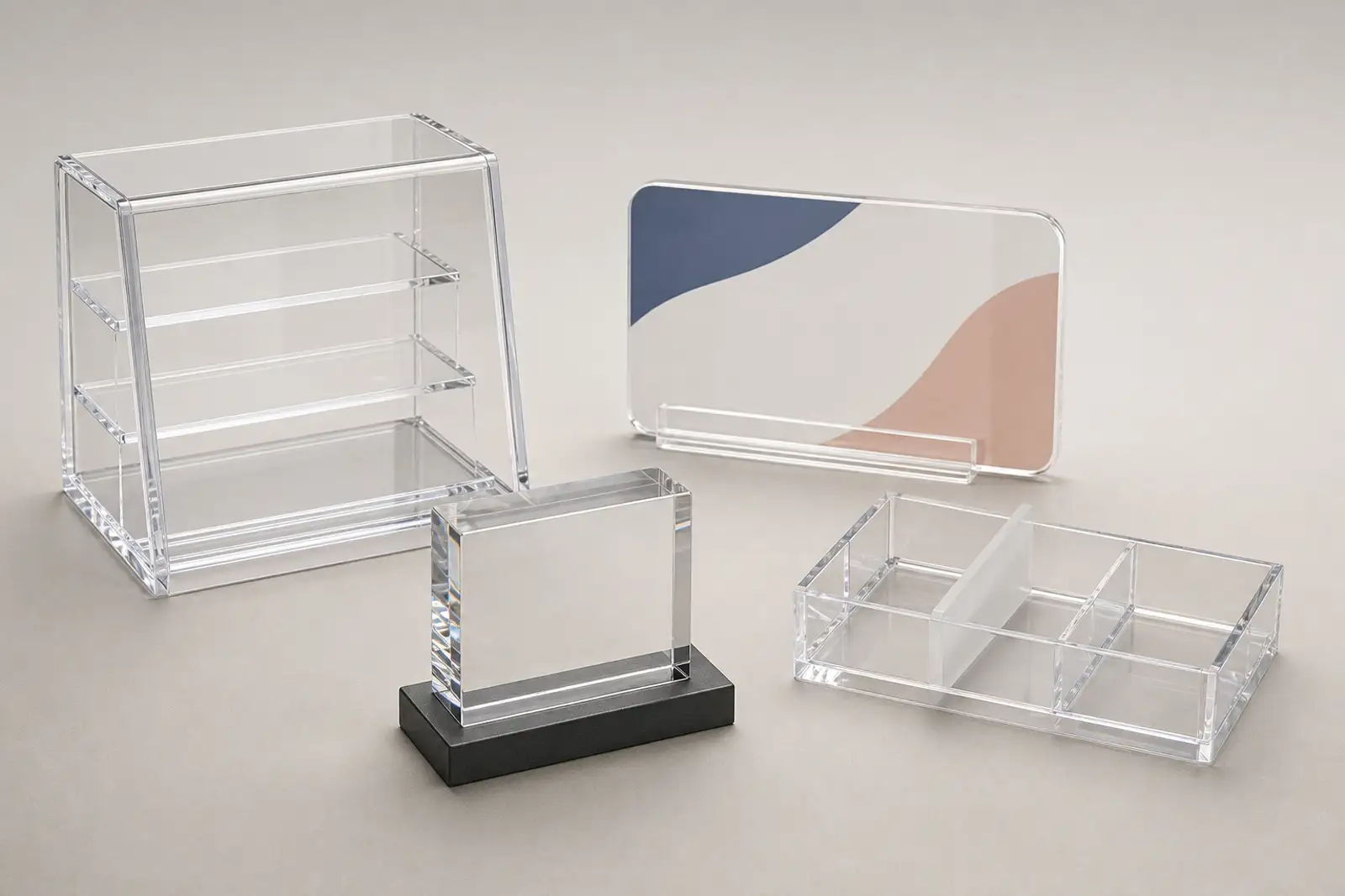 Range of finished custom acrylic fabrication products on a neutral studio surface; a clear acrylic POP display case, a UV-printed brand sign, a polished acrylic award block, and a cosmetics organizer with frosted dividers